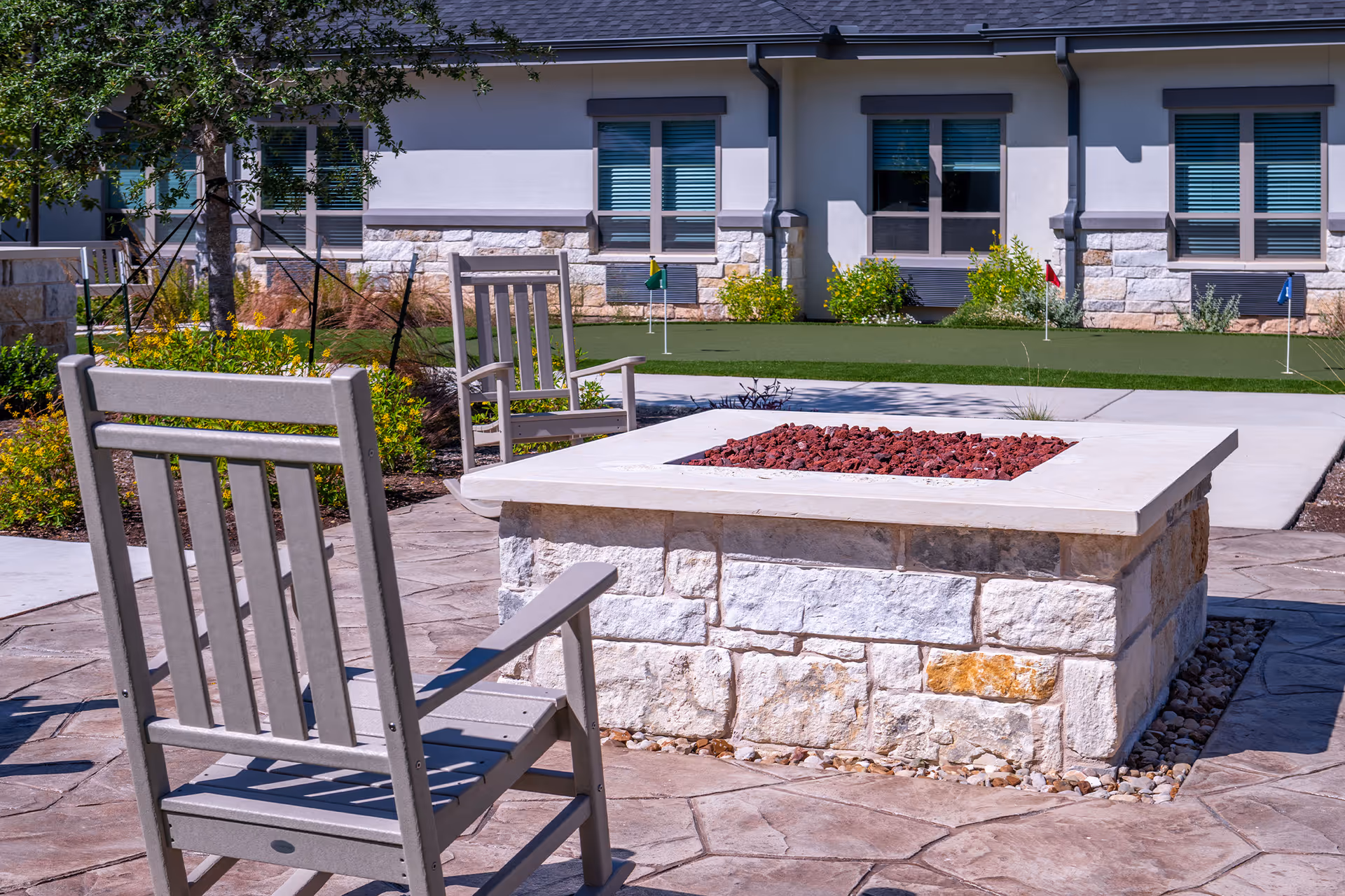 Outdoor courtyard with a stone fire pit, wooden rocking chairs, and a putting green in front of the senior living building.