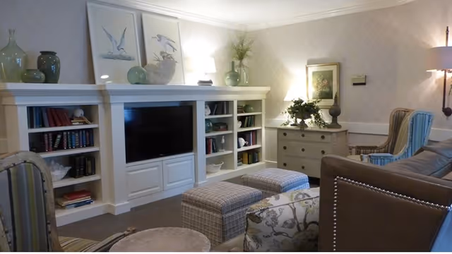 A cozy living room with a built-in white entertainment center housing a flat-screen TV and shelves filled with books and decorative items. The room features patterned wallpaper, a chest of drawers with plants and a framed picture on top, a leather sofa, upholstered chairs, two plaid ottomans, and soft lighting from a wall lamp and table lamp.