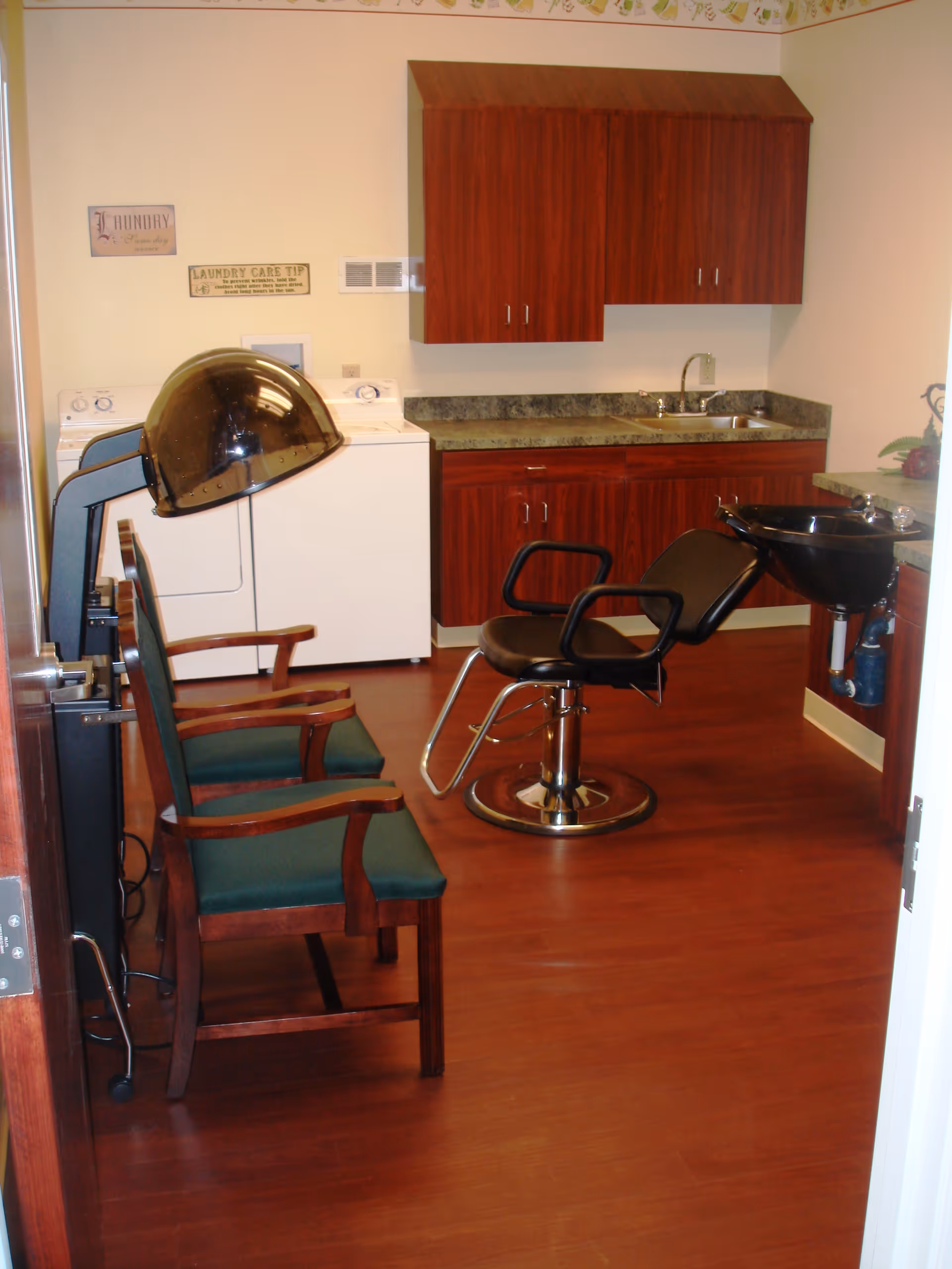 Interior room with a salon styling chair and hood dryer, waiting chairs, sink and cabinetry beside a washer and dryer.