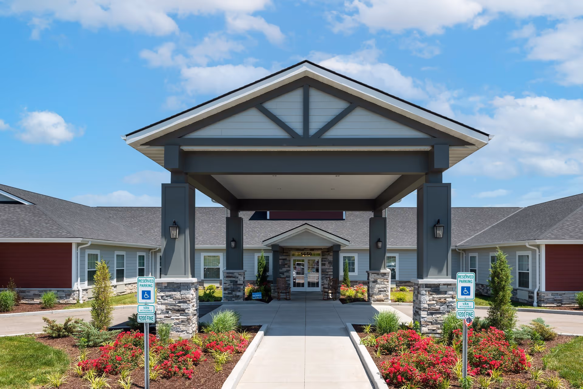 Front entrance of Oak Pointe of Washington senior living facility with a covered driveway, stone pillars, landscaped flower beds with red flowers, and a clear blue sky.