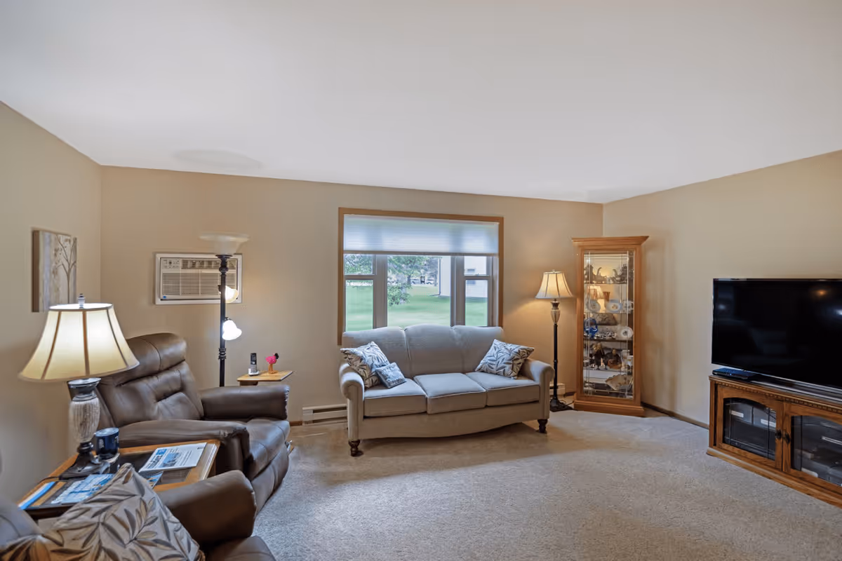 A cozy living room with beige walls and carpet, featuring a gray sofa with patterned pillows under a window, two brown leather recliners with a side table and lamp, a wooden display cabinet with decorative items, and a flat-screen TV on a wooden stand.