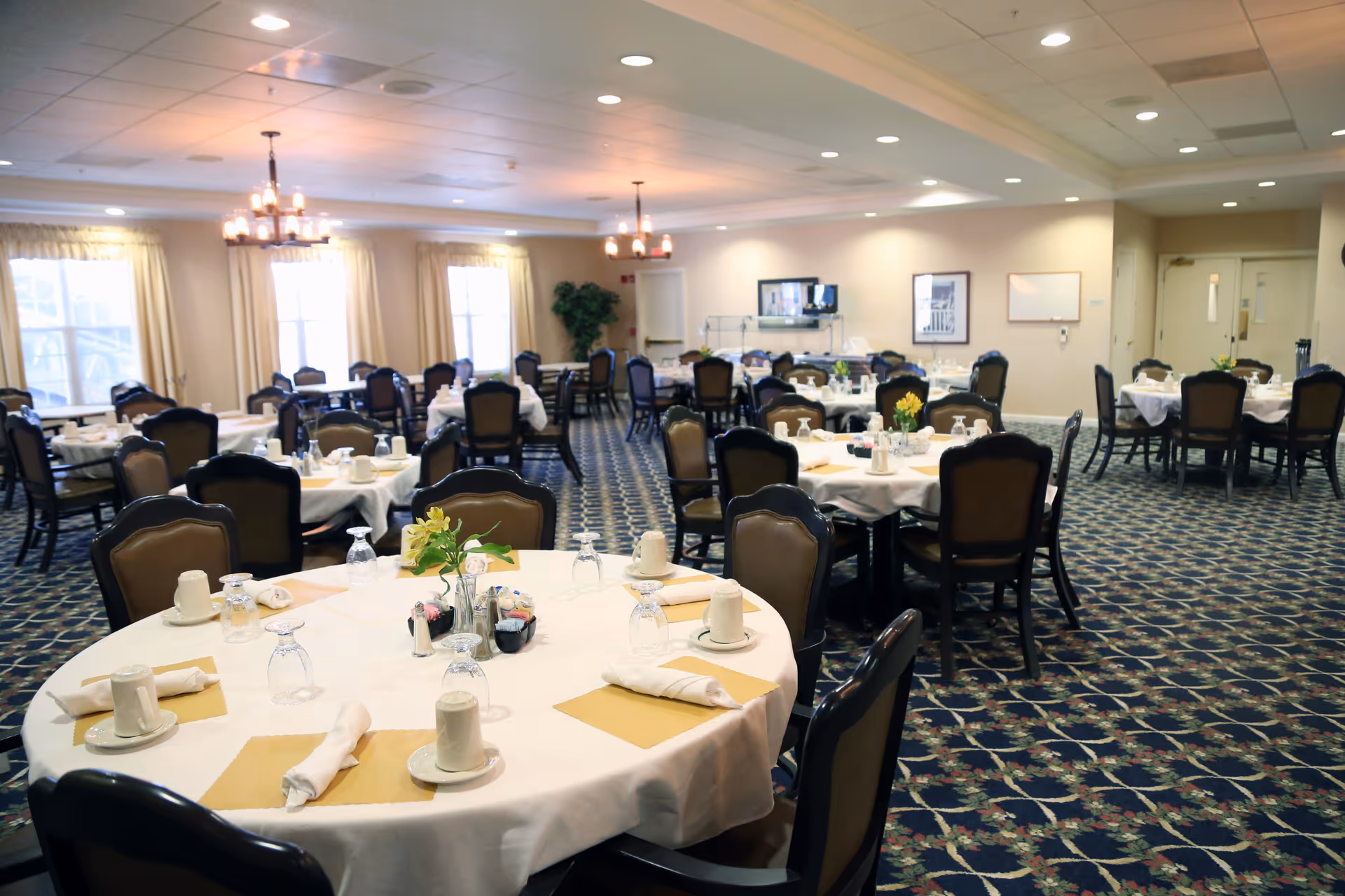 A spacious dining room with multiple round tables covered with white tablecloths, each set with cups, glasses, napkins, and small flower centerpieces. The room has patterned carpet flooring, beige walls, large windows with curtains, and ceiling lights including chandeliers.