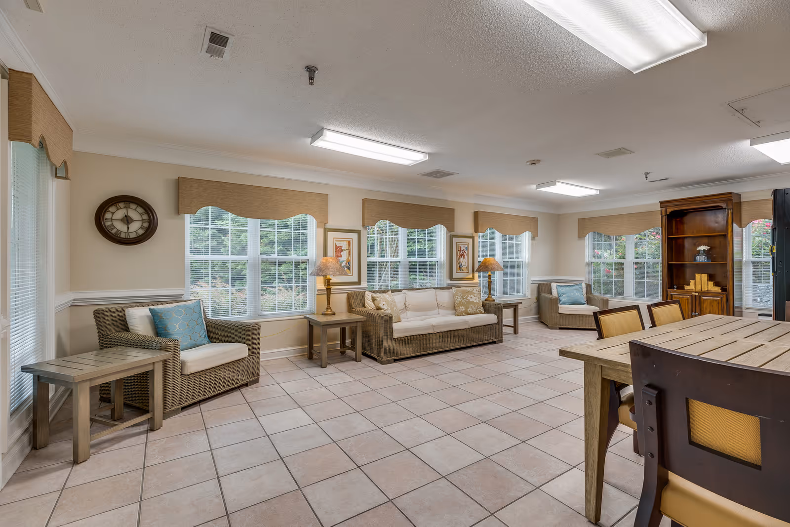 A bright and spacious living room in an assisted living facility featuring wicker furniture with cushions, including a sofa and armchairs, side tables with lamps, a wooden dining table with chairs, large windows with beige valances, a wall clock, framed artwork, and a wooden cabinet with decorative items.