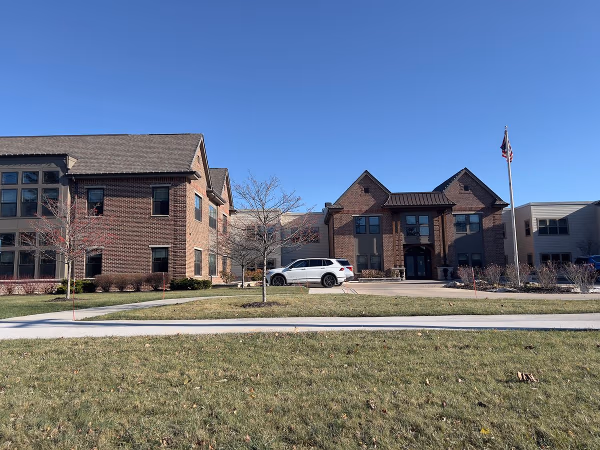 Brick multi-story assisted living building with a white SUV parked at the entrance, an American flag on a pole, and a grassy lawn under a clear blue sky.
