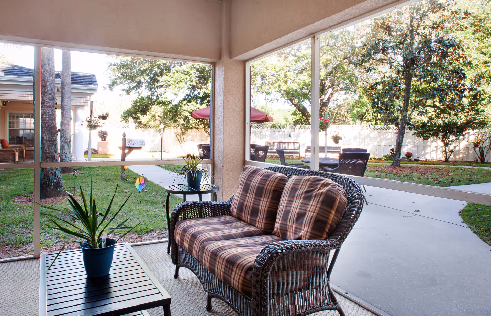 A screened-in porch area with a wicker loveseat featuring plaid cushions, a small round side table with a potted plant, and a rectangular coffee table with another potted plant. Outside the porch, there is a garden with green grass, trees, a white fence, and outdoor seating with umbrellas.