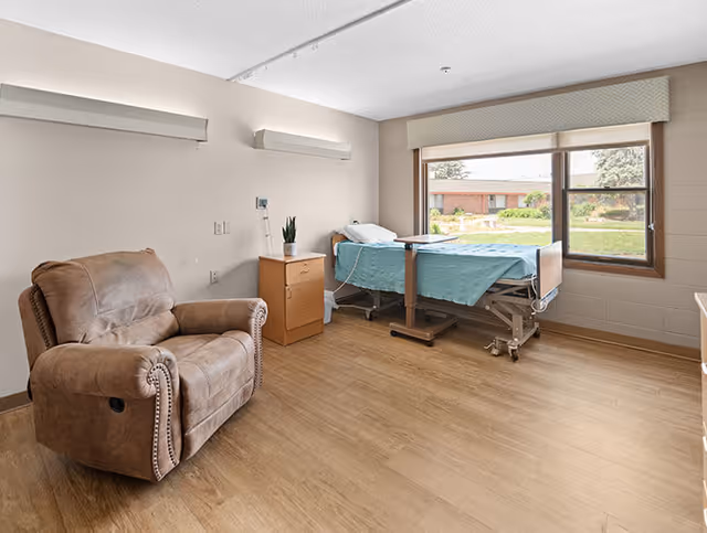 A senior living facility room with a hospital-style bed covered with a light blue blanket positioned near a large window showing an outdoor garden. Next to the bed is a wooden nightstand with a small plant on top. A comfortable brown recliner chair is placed on the left side of the room. The floor is wooden, and the walls are painted beige.