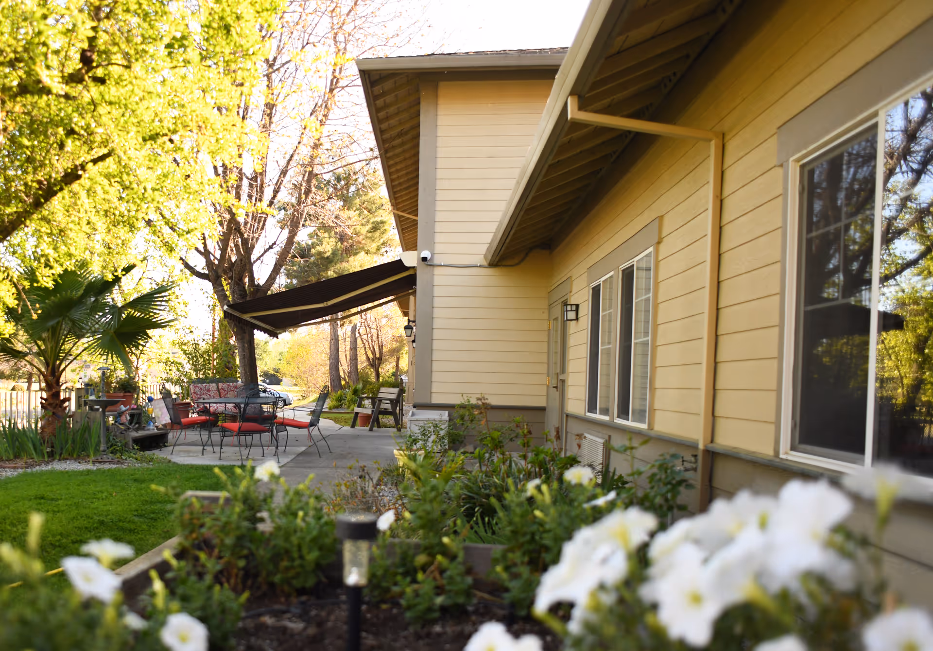 Outdoor patio area at Oakdale Heights Assisted Living featuring a yellow building with windows, a concrete patio with metal chairs and a table with red cushions, surrounded by green plants and trees in a sunny setting.