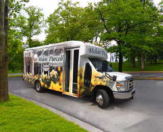 A white shuttle bus with floral graphics and the text 'Helen Purcell' parked on a paved road surrounded by green trees and grass.