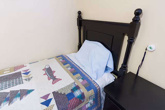 Single twin bed with a black wooden headboard, patterned quilt, blue pillow, and a black nightstand against a beige wall.