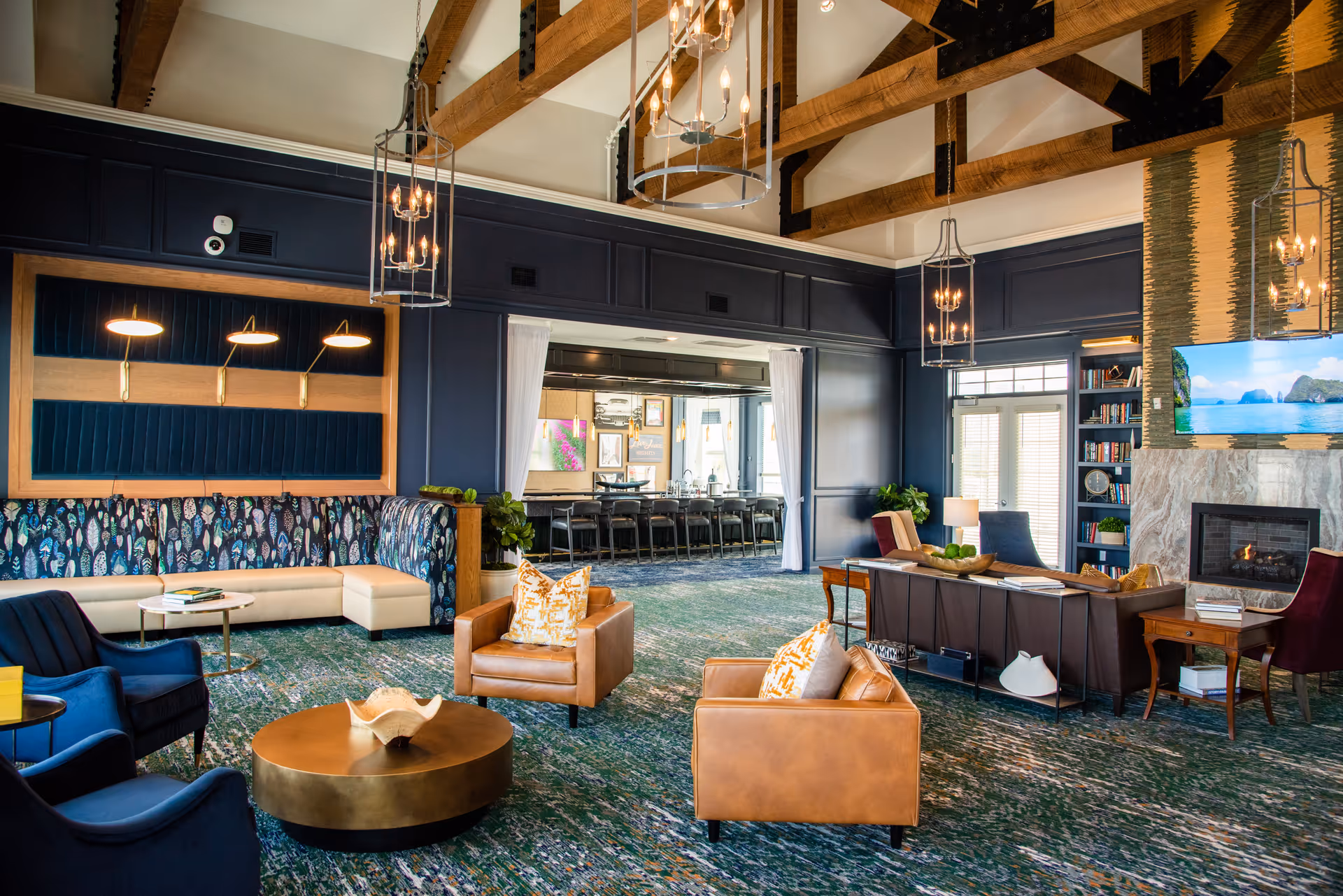 Open, upscale communal lounge with leather armchairs, blue seating, a round coffee table, fireplace, built-in shelving and vaulted beamed ceiling.