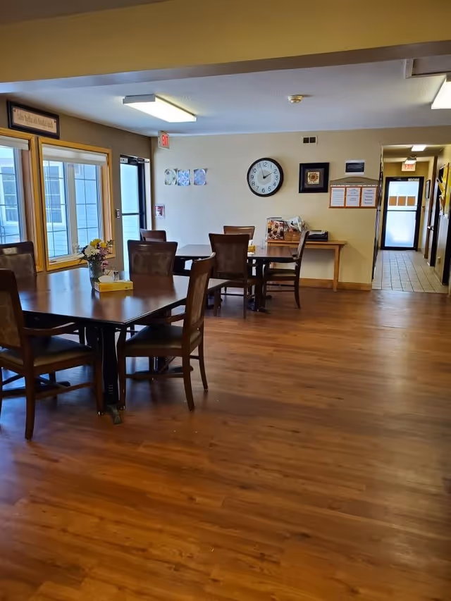 Interior view of a dining area with wooden floors, several wooden tables and chairs, a vase with flowers on one table, a clock on the wall, and a hallway leading to a door in the background.