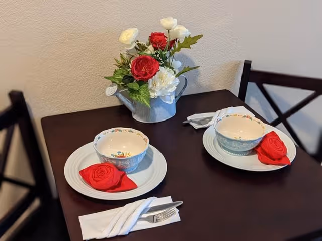 A small dark wooden dining table set for two with white plates, floral patterned bowls, and red napkins folded into rose shapes. A white napkin with a fork and knife is placed beside one setting. A metal watering can filled with red and white artificial flowers serves as a centerpiece. Two dark wooden chairs with cross-back design are partially visible.