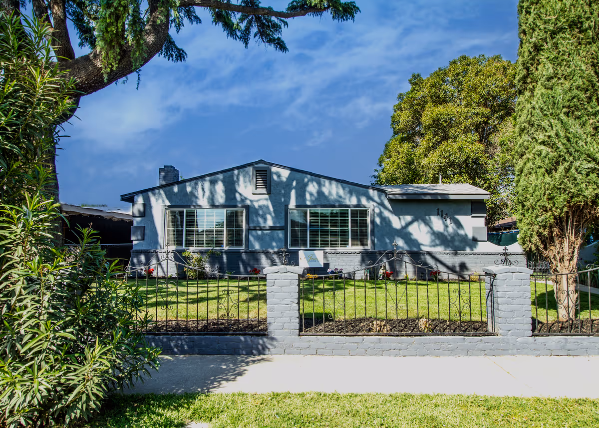 Front exterior view of a single-story residential building with two large windows, a small lawn, and a decorative metal fence. The building is painted gray with darker gray trim and surrounded by trees and shrubs under a blue sky.