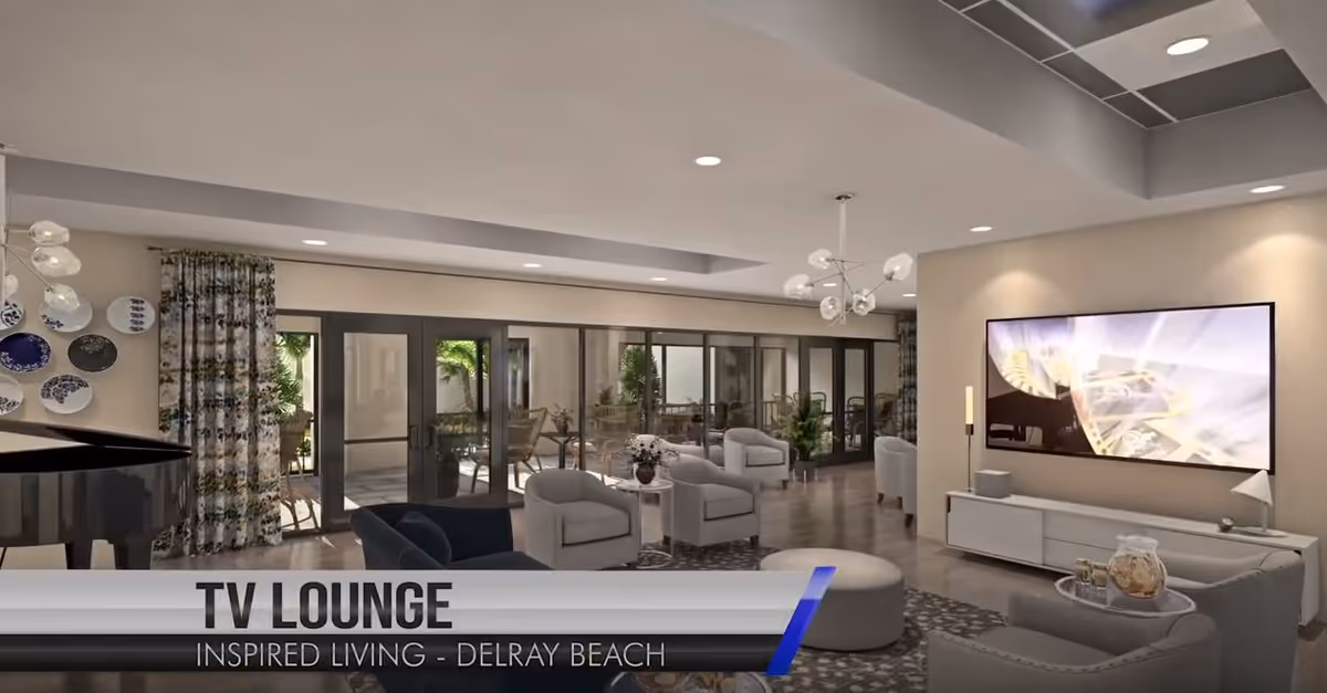 A modern TV lounge with comfortable seating including armchairs and sofas arranged around a large flat-screen TV mounted on the wall. The room features large glass doors leading to an outdoor patio area with greenery visible outside. Decorative plates are displayed on one wall, and a grand piano is partially visible on the left side. The space is well-lit with ceiling lights and a contemporary chandelier.