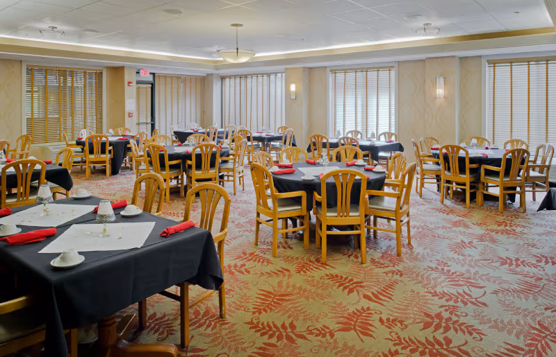 A spacious dining room with multiple round and square tables covered with black tablecloths and white placemats. Each table is set with cups, saucers, and red napkins. The room has wooden chairs, large windows with blinds, patterned carpet with red and beige tones, and soft lighting from ceiling fixtures and wall sconces.