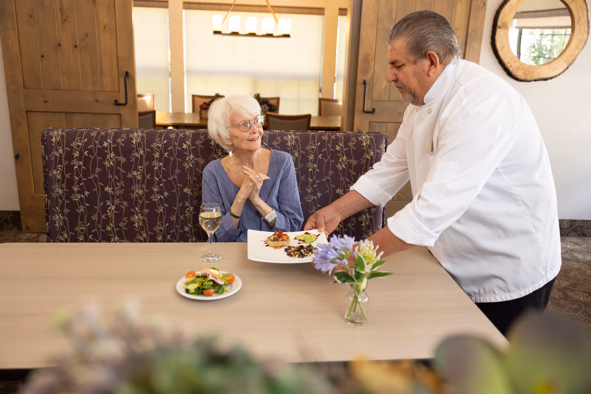 An elderly woman with white hair and glasses sitting at a dining table with a glass of white wine, smiling and looking at a chef who is serving a plate of food to her in a cozy dining room with wooden doors and a floral-patterned bench.