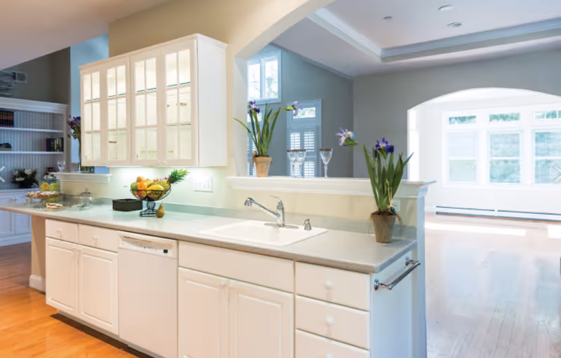 Bright open kitchen with white cabinets, a sink and countertop island overlooking a sunny adjoining living area.