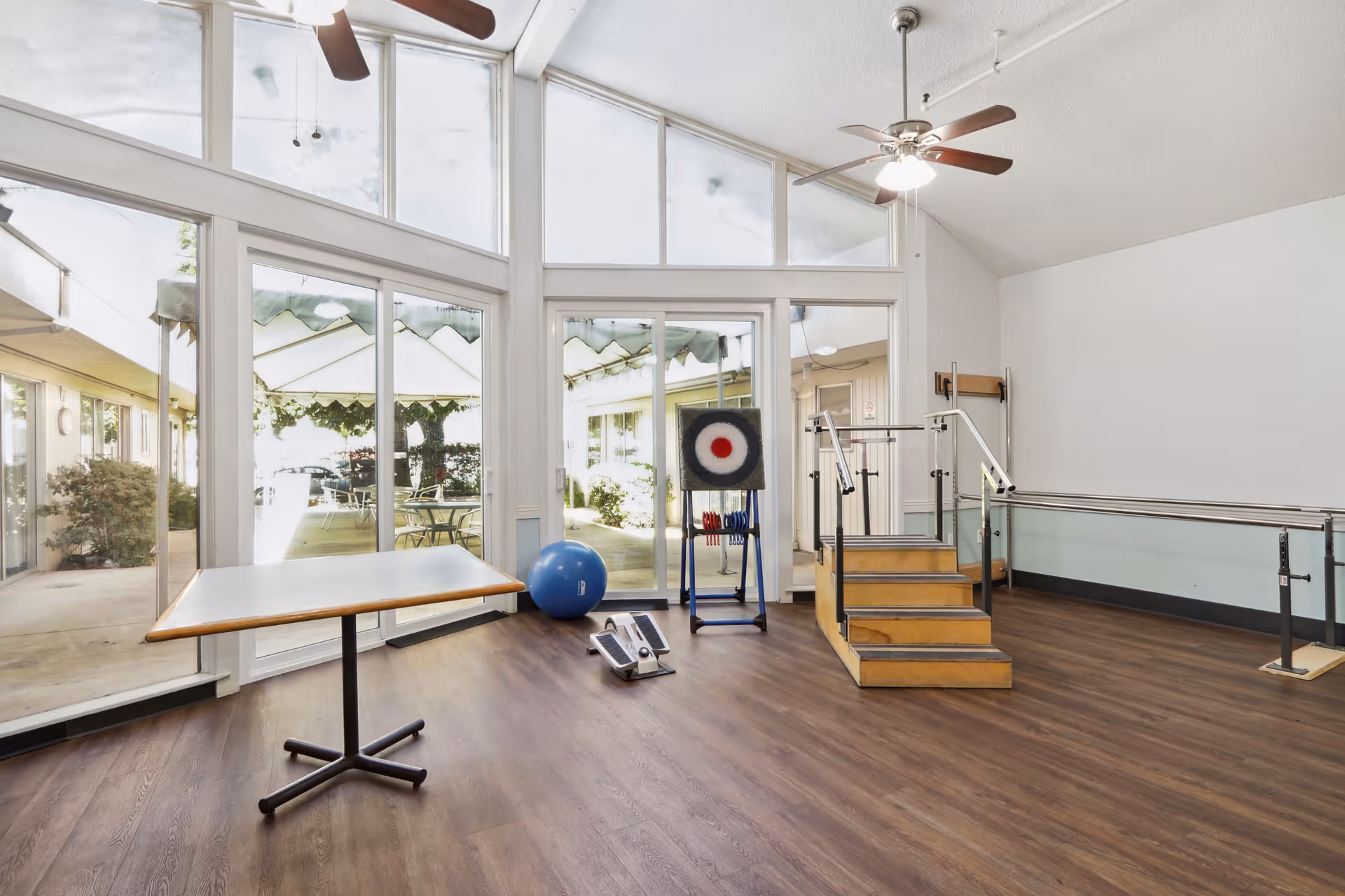 A bright room with large windows and glass doors leading to an outdoor patio area. Inside the room, there is a small wooden table, a blue exercise ball, a stepper exercise machine, a target board on a stand, and a set of wooden stairs with handrails for physical therapy or rehabilitation. The room has wooden flooring, ceiling fans, and white walls.