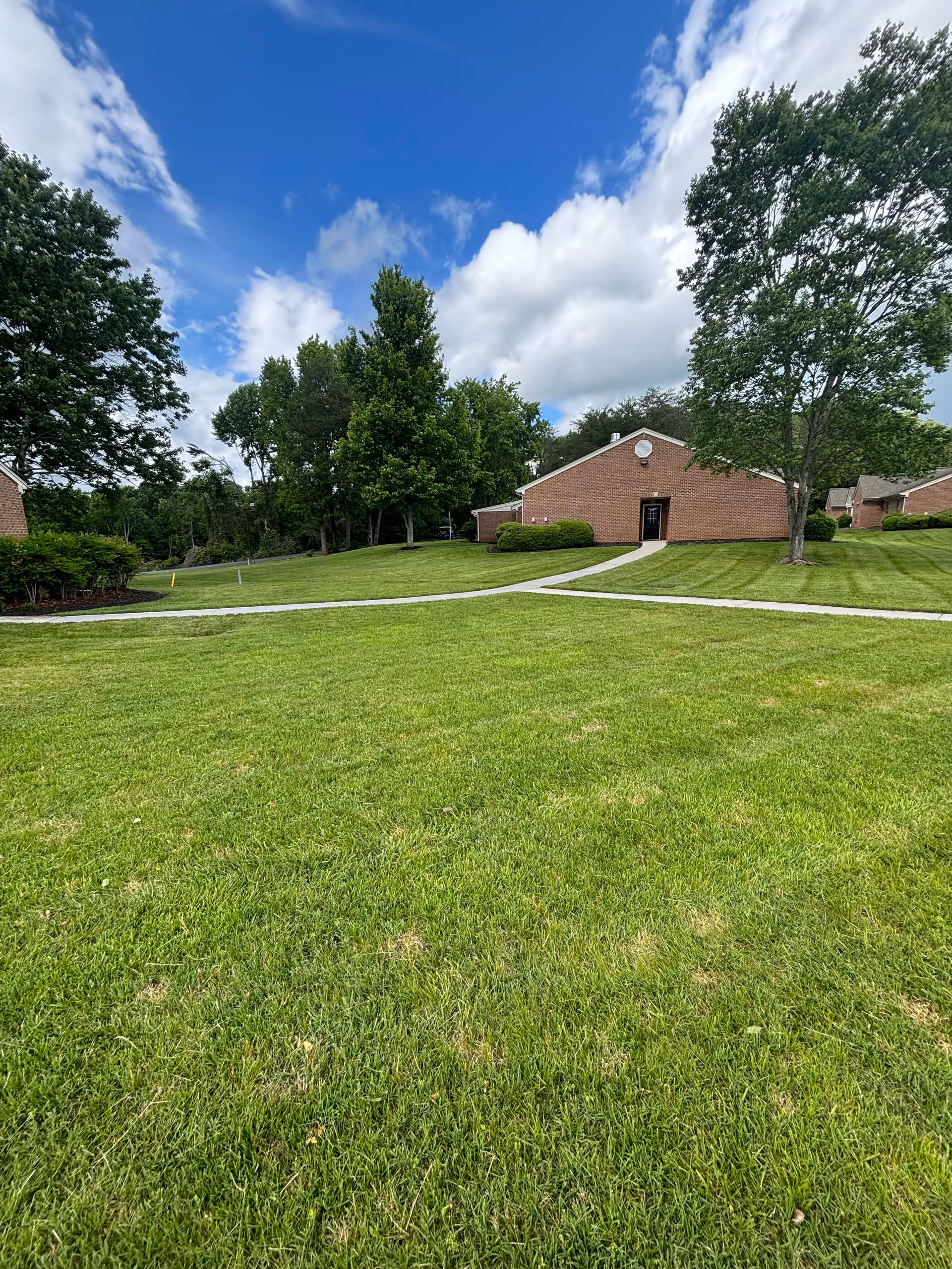 A well-maintained grassy lawn with a concrete walkway leading to a single-story brick building surrounded by trees under a partly cloudy blue sky.