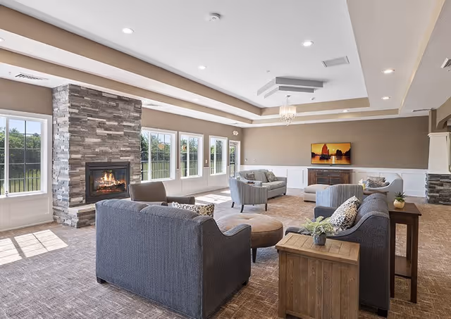 A spacious and well-lit living room with large windows allowing natural light to fill the space. The room features a stone fireplace with a fire burning, multiple gray upholstered sofas and armchairs arranged around a central ottoman, a wooden side table with a small plant, and a flat-screen TV mounted on the far wall. The ceiling has recessed lighting and a decorative chandelier, with beige walls and carpeted flooring.