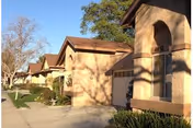 Exterior view of a residential-style building with beige stucco walls and brown roofs, surrounded by trees and shrubs, under a clear blue sky.