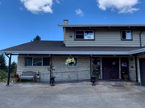 Exterior view of a two-story building with a stone and siding facade under a blue sky with some clouds. The entrance features a covered porch with two large planters, a bench, and a double door with a wreath. There are windows on both floors and some potted plants near the entrance.