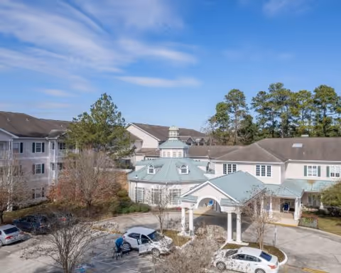 Exterior view of King's Preserve at Kingwood senior living facility showing a multi-story building with a light green roof, a covered entrance with columns, several parked cars, trees, and a clear blue sky.