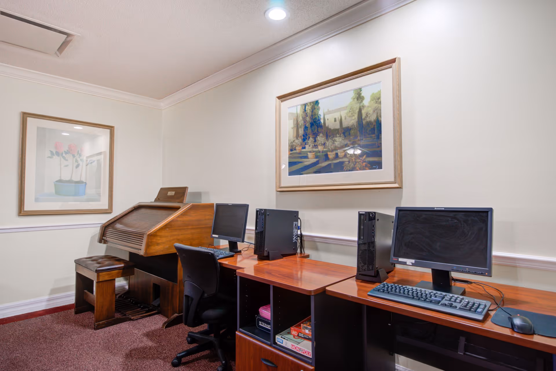 A small room with two desktop computers on wooden desks, each with a monitor, keyboard, and mouse. There is a black office chair in front of one desk. To the left, there is a wooden roll-top desk with a matching bench. The walls are light-colored and decorated with two framed paintings, one of flowers and the other of a garden scene. The floor is carpeted in a dark red color.