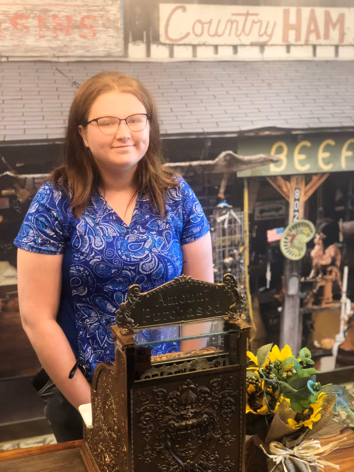 A young woman with glasses and a blue patterned shirt stands behind an ornate antique cash register on a wooden counter. To the right of the cash register is a bouquet of sunflowers wrapped in burlap. The background features a rustic storefront with signs that read 'Country HAM' and 'BEEF'.