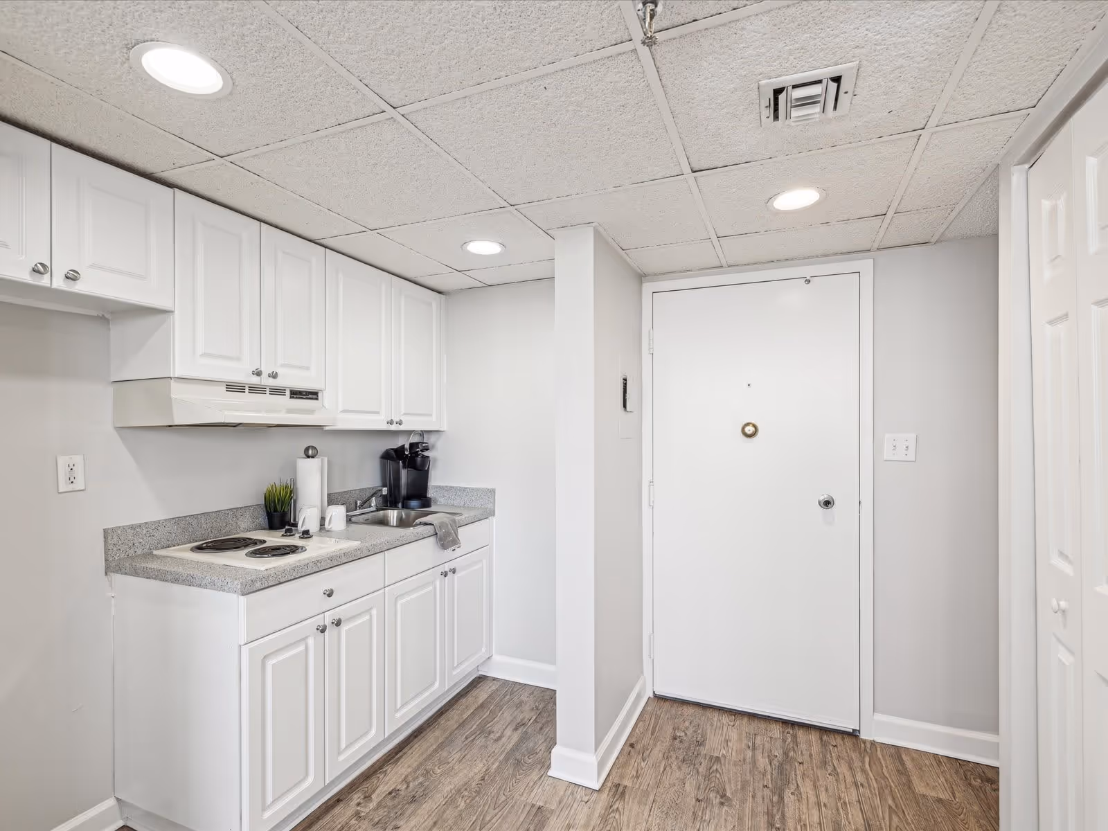 Small kitchen area with white cabinets, a countertop with a sink, a coffee maker, and a two-burner electric stove. The kitchen is adjacent to a white door and has wood-style flooring and a drop ceiling with recessed lighting.