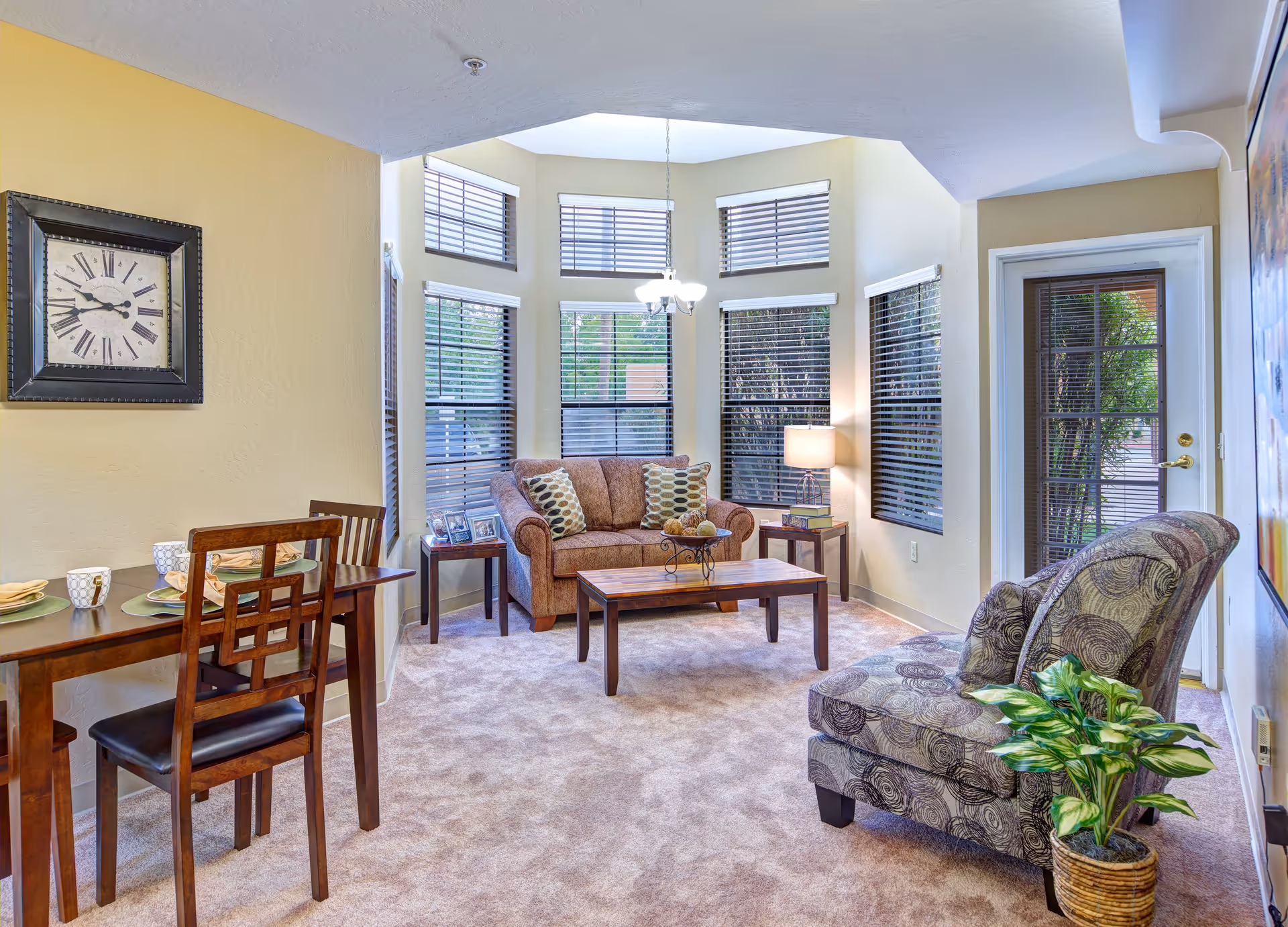 A cozy living room area with beige walls and carpet. The room features a brown loveseat with patterned pillows, two side tables with a lamp and photo frames, a wooden coffee table, and two patterned armchairs next to a potted plant. There is a dining table with two chairs and place settings on the left side. Multiple windows with blinds allow natural light to enter, and a glass door leads outside.