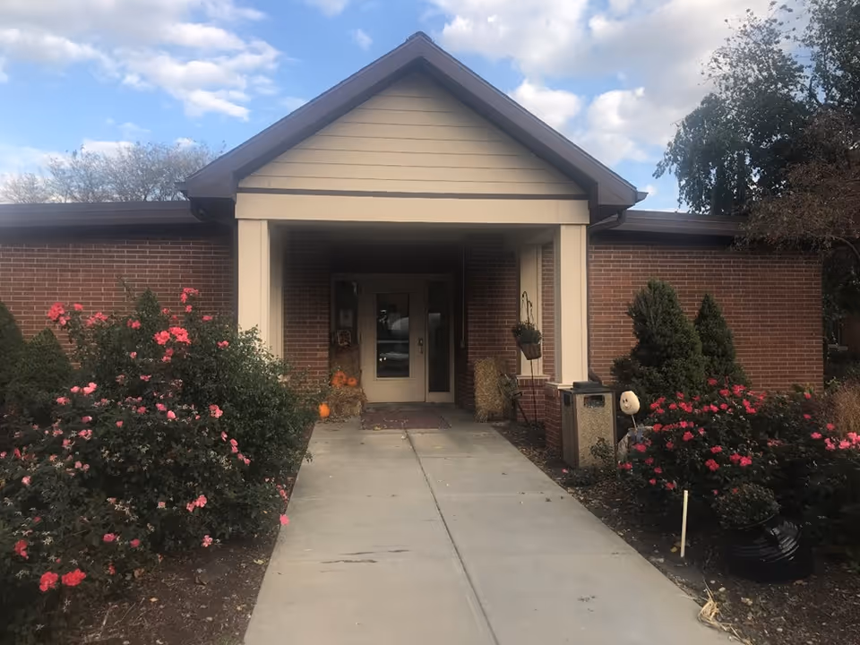 Front entrance of a single-story brick care facility with a covered porch, walkway, and flowering shrubs.