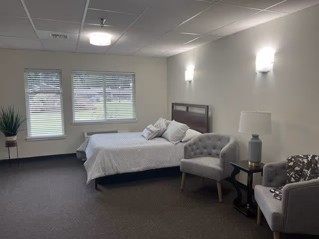 Furnished bedroom with a bed, two armchairs, side table and window with blinds in a senior living facility.
