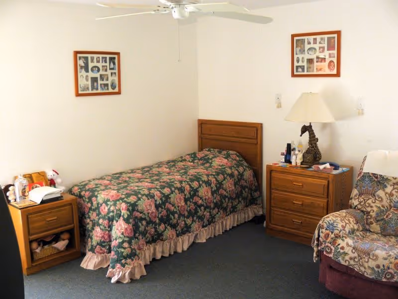 A small bedroom with a single bed covered in a floral bedspread. There are two wooden nightstands on either side of the bed, one with a lamp shaped like a horse and various personal items. A floral-patterned armchair is positioned next to one nightstand. Two framed photo collages hang on the white walls above the bed.