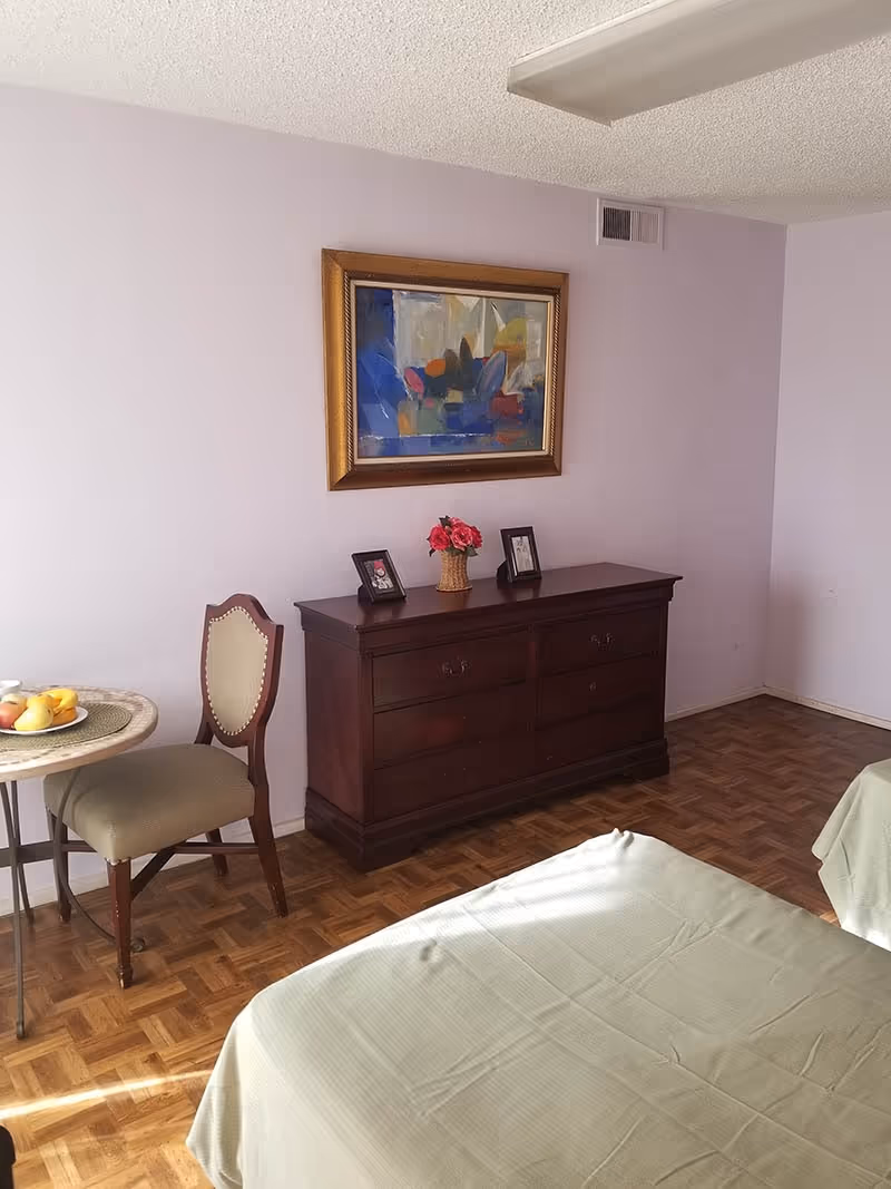 A bedroom with two beds covered in light green bedspreads, a wooden dresser with a vase of pink flowers and two framed photos on top, a small round table with a chair, and a colorful abstract painting hanging on a light purple wall.