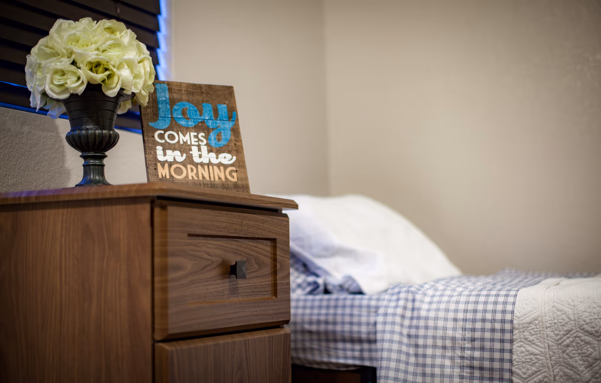 Wooden nightstand topped with a vase of white flowers and a sign that reads "Joy comes in the morning" beside a neatly made bed with blue plaid bedding.