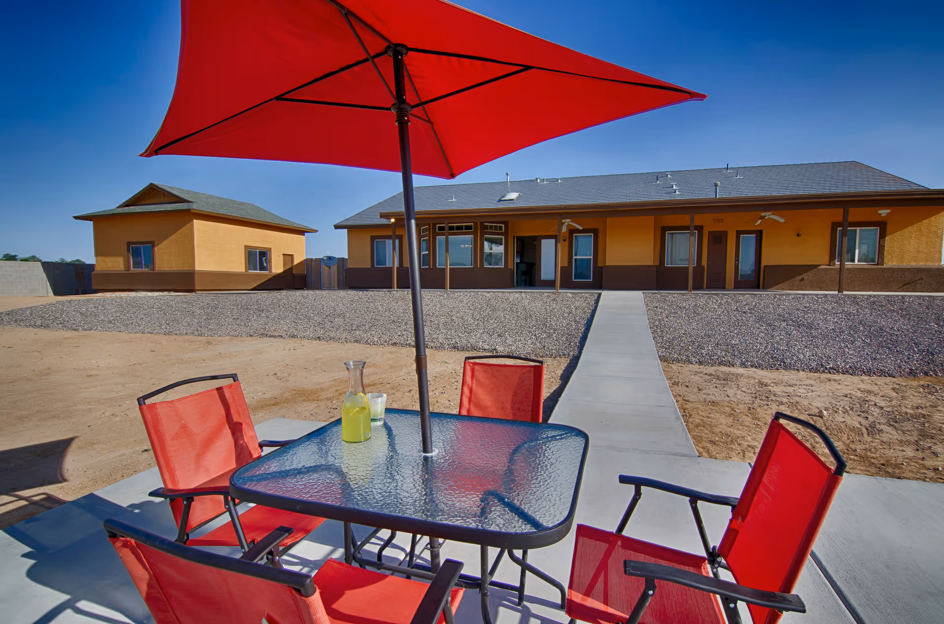 Outdoor patio area with a glass table and four red chairs under a large red umbrella. A pitcher of lemonade and a glass are on the table. In the background, there is a single-story building with a beige and brown exterior and a clear blue sky.