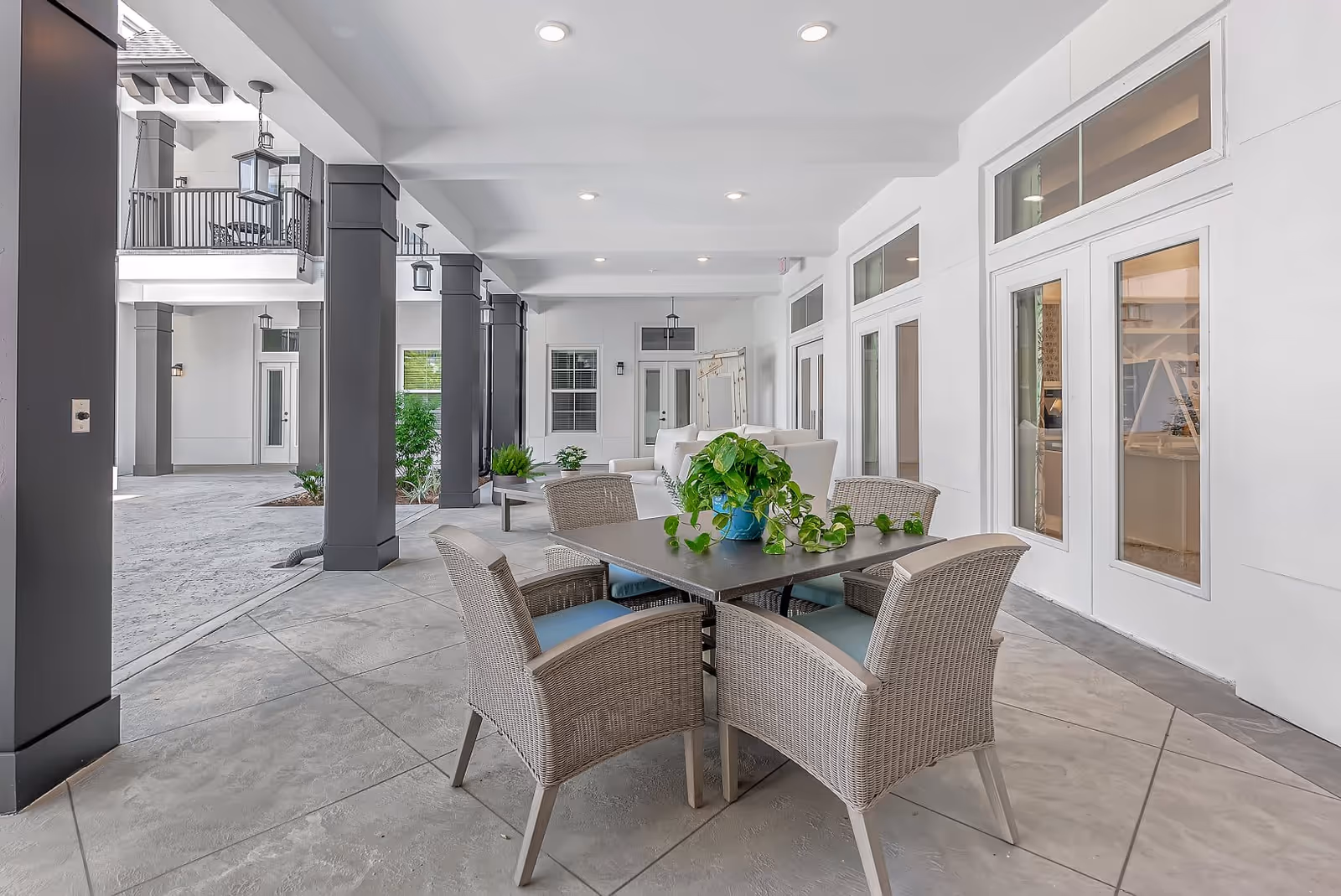 Covered outdoor seating area at The Blake at Miramar Beach featuring a square table with four wicker chairs with blue cushions, a potted green plant on the table, white walls with multiple glass doors and windows, black square columns, and additional seating with white sofas in the background.