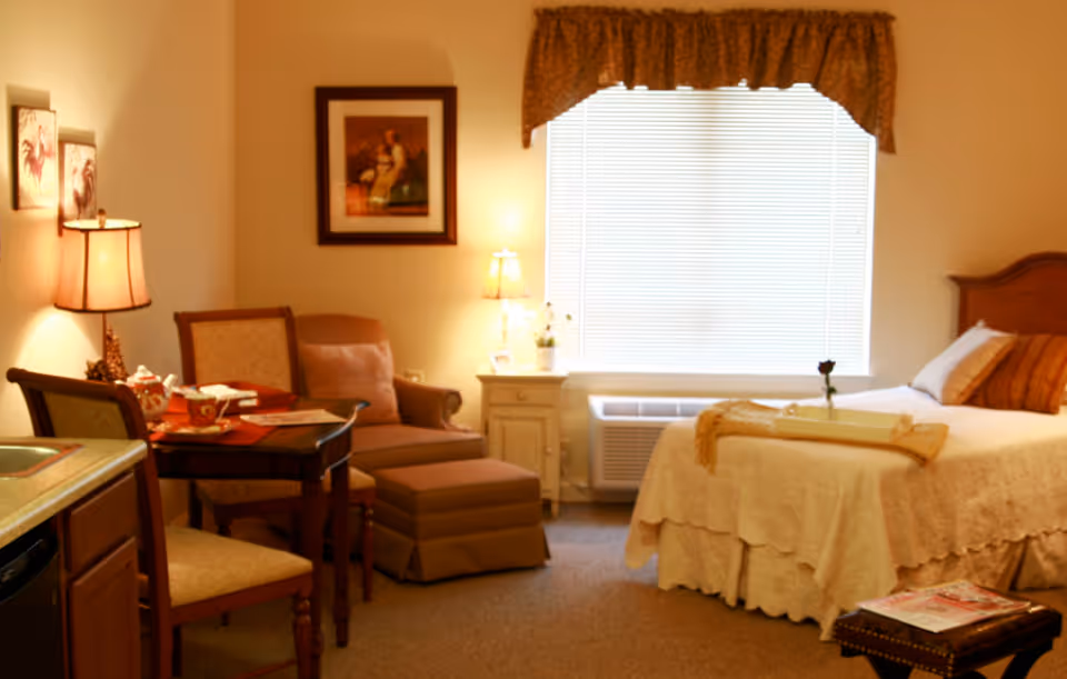 A cozy senior living facility room with a single bed covered in a cream-colored bedspread and a brown pillow. Next to the bed is a small nightstand with a lamp and a small plant. A window with blinds and a valance is behind the nightstand. There is a comfortable armchair with an ottoman and a side table with a lamp and framed artwork on the wall. A small dining table with two chairs is partially visible, along with a kitchenette area on the left side.