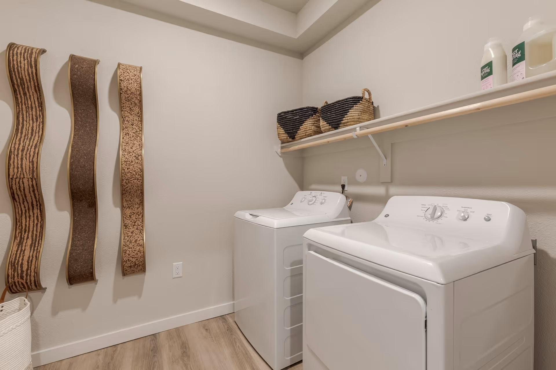 Laundry room with a washer and dryer under a shelf holding baskets and detergent and decorative wall hangings.