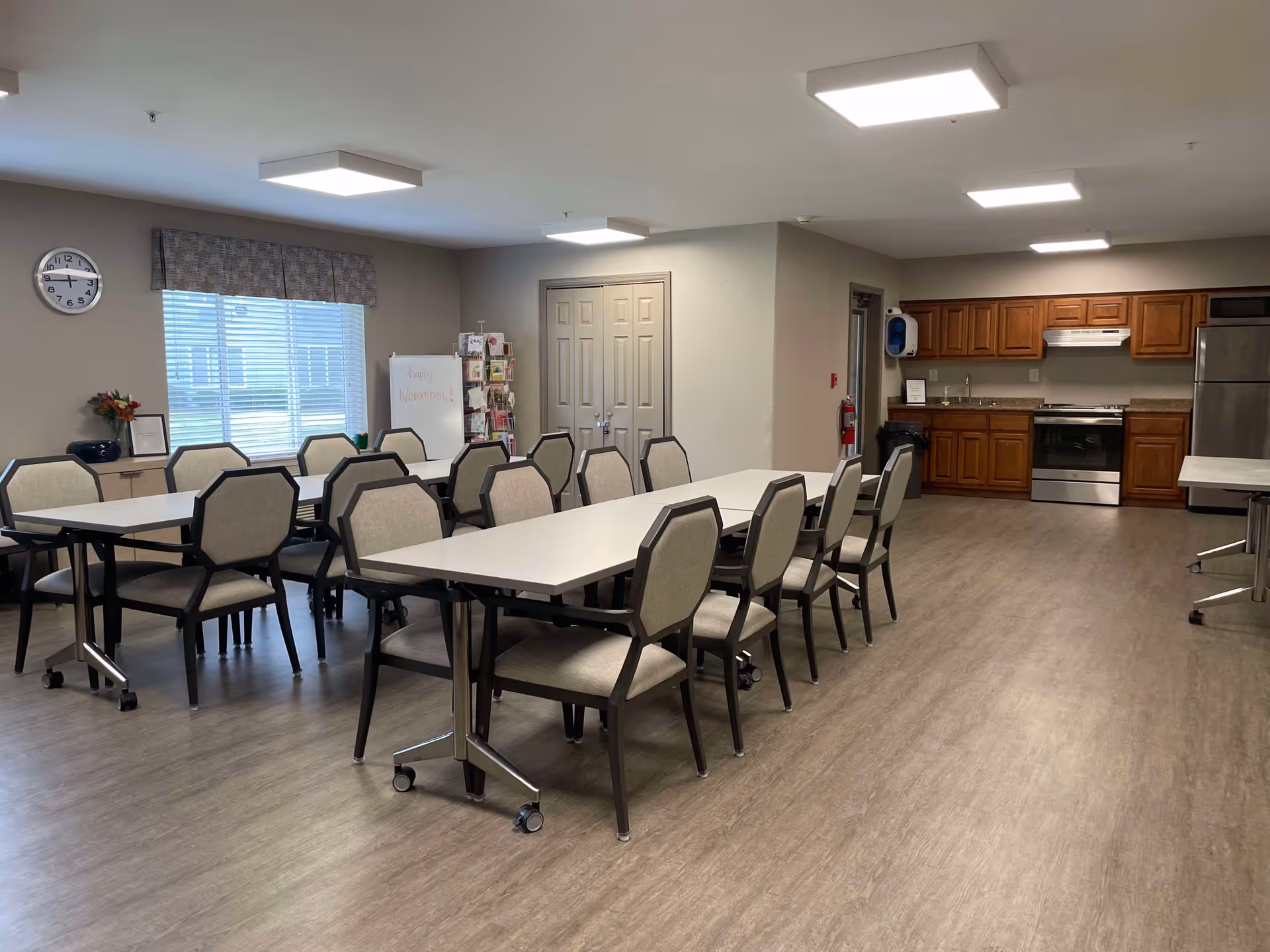 A spacious dining room with multiple tables and chairs arranged neatly. The room has a large window with blinds and a clock on the wall. In the background, there is a kitchen area with wooden cabinets, a stove, and a refrigerator. The floor is covered with wood-like flooring, and the ceiling has rectangular light fixtures.
