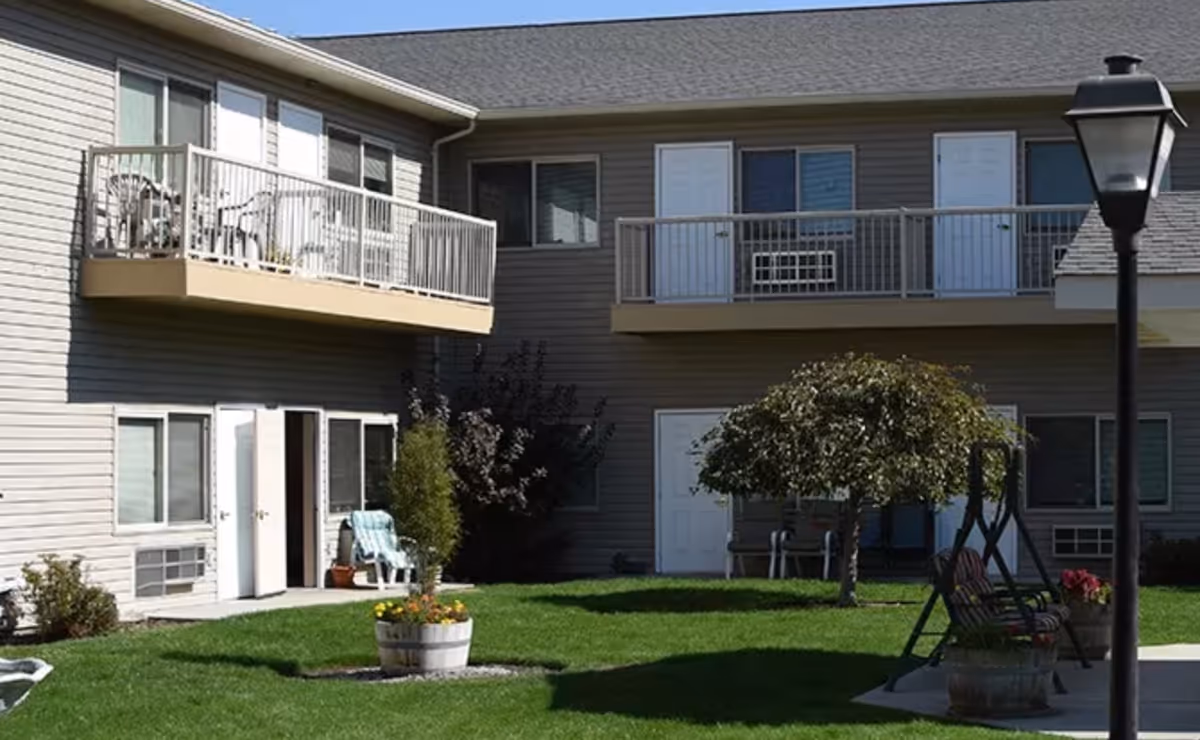 Courtyard of a two-story residential building with balconies, a grassy lawn, potted flowers, patio chairs, a small tree, and a lamppost.