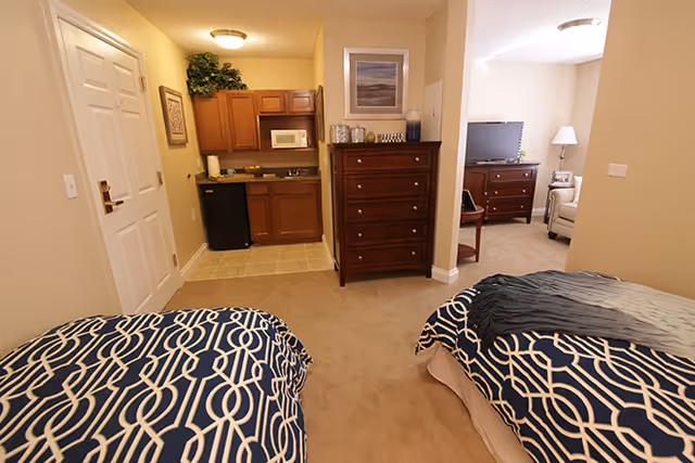 Interior view of a senior living facility room with two twin beds covered in blue and white patterned bedding. The room has beige walls and carpet. There is a wooden dresser with a framed picture above it, and a small kitchenette area with cabinets, a microwave, and a mini fridge near the entrance door. In the background, there is a sitting area with a TV on a wooden stand and a beige armchair with a floor lamp beside it.