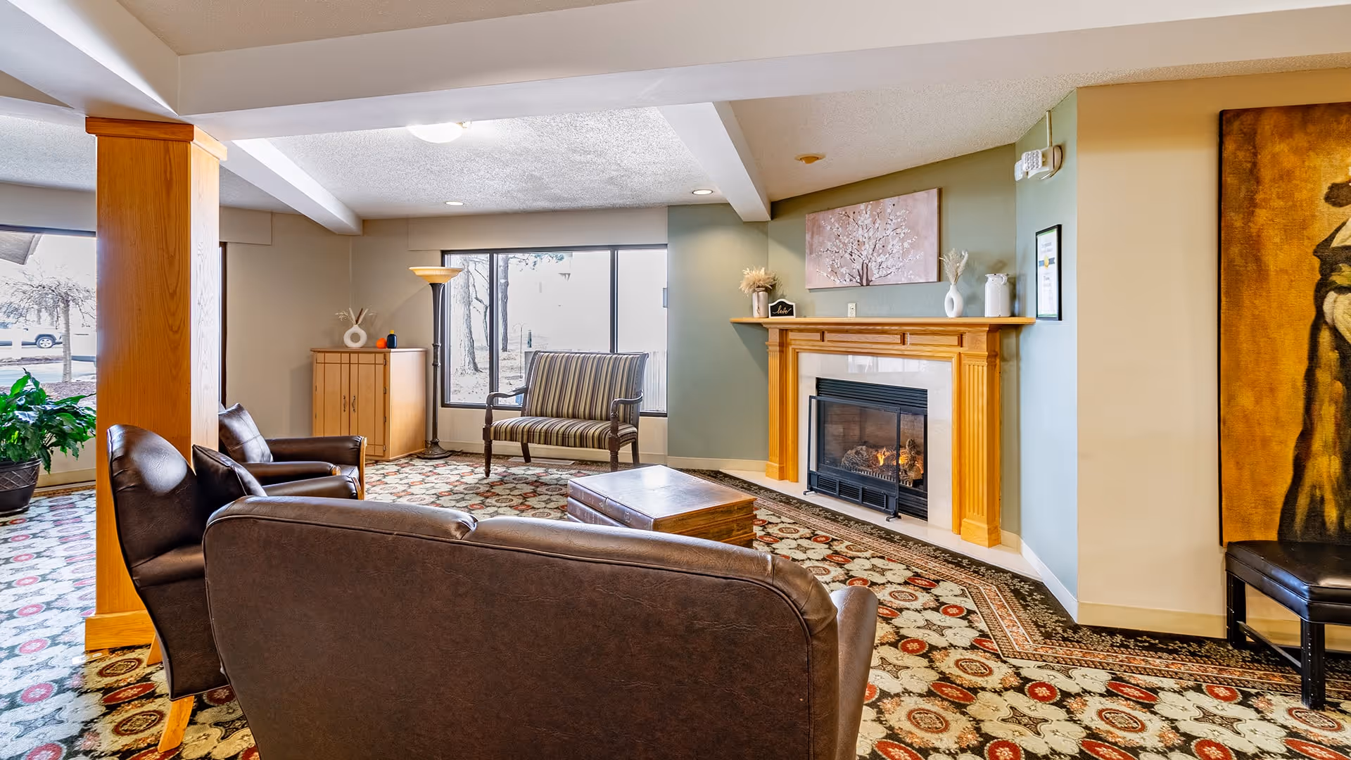 A cozy living room area with a brown leather sofa and armchairs arranged around a wooden coffee table. There is a fireplace with a wooden mantel on the right wall, decorated with vases and a framed picture above it. A striped loveseat is positioned near large windows letting in natural light. The room features patterned carpet flooring and light-colored walls with wooden accents.