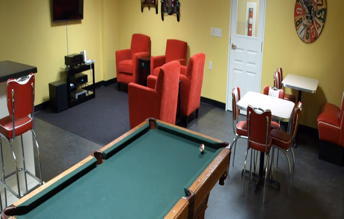 Recreation/lounge room with a pool table in the foreground, red upholstered chairs, diner-style tables and chairs, and a wall-mounted TV.