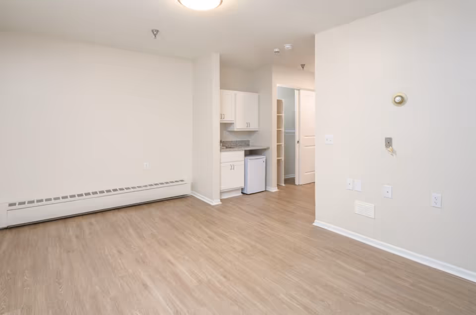 Empty studio-style living area with light wood flooring, neutral walls, and a small kitchenette with cabinets and a mini fridge.