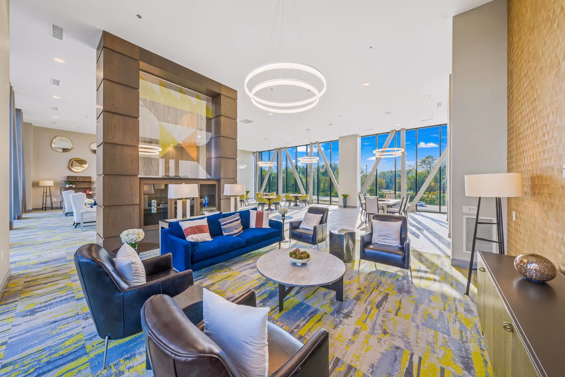 A bright and spacious living room area in a senior living facility with large floor-to-ceiling windows letting in natural light. The room features a blue sofa, several black leather armchairs with white pillows, a round coffee table with decorative items, modern circular ceiling lights, and a large wooden partition with an abstract art piece. The carpet has a colorful pattern in shades of blue, yellow, and gray. In the background, there are additional seating areas and dining tables near the windows.
