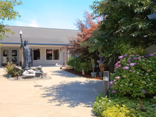 Outdoor courtyard area of a senior living facility with a small statue resembling the Statue of Liberty, surrounded by plants, flowers, and trees. The building with windows and a door is visible in the background under a clear blue sky.