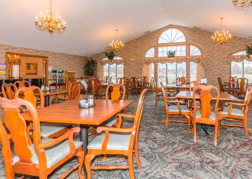 A spacious dining room with multiple wooden tables and chairs arranged neatly. The room features large windows with decorative curtains, allowing natural light to brighten the space. Elegant chandeliers hang from the ceiling, and there are plants placed near the windows and walls. The carpet has a patterned design, and the walls are covered with a textured wallpaper.