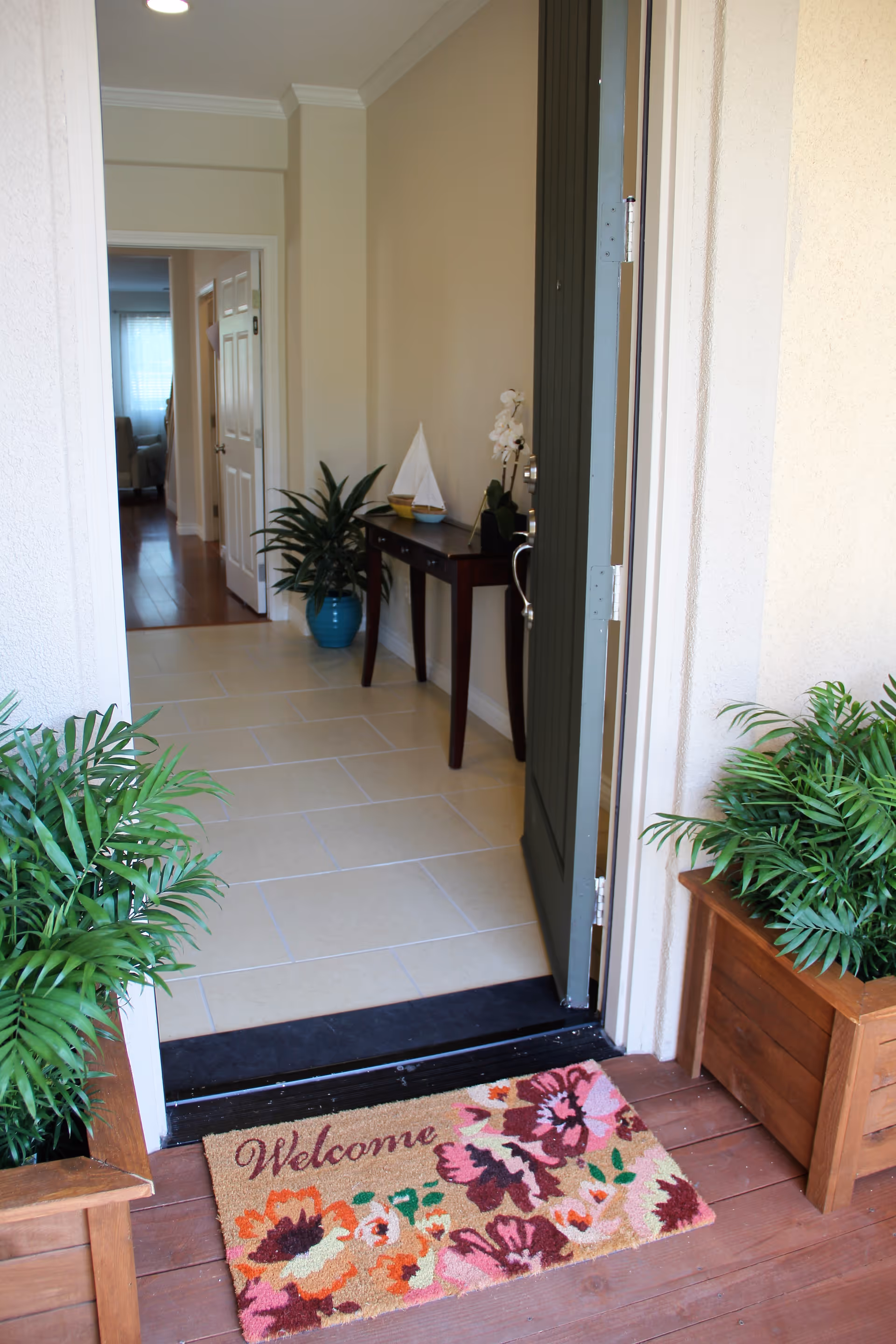 Open front door showing an entryway with a welcome mat featuring colorful flowers. Two potted plants flank the doorway on a wooden floor. Inside, there is a tiled floor, a small table with decorative items including a small sailboat model and a potted orchid, and a hallway leading to other rooms with wooden flooring.